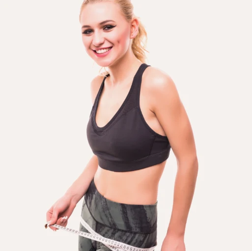 A woman wearing a black sports bra and grey patterned leggings, measuring her waist with a white tape measure